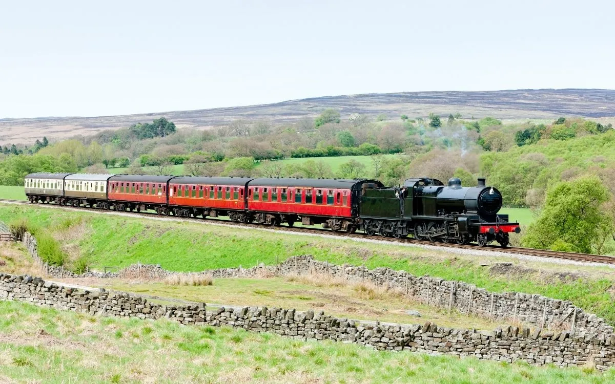 Ein Dampfzug der North Yorkshire Moors Railway fährt an einer Trockenmauer vorbei durch die Heidelandschaft